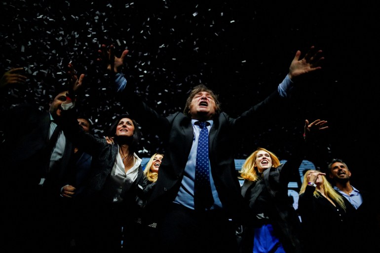 During a campaign rally, Javier Milei lifts both arms in the air, in a gesture of seeming ecstasy. Confetti appears to rain down in the darkened room, filled with supporters.