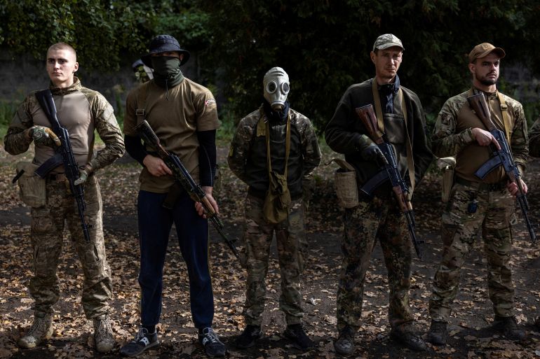 A group of five volunteers posing for a photo. They are doing basic training for the Ukraine armed forces. One of the men has covered his face. One is wearing a gas mask. They are all holding weapons.