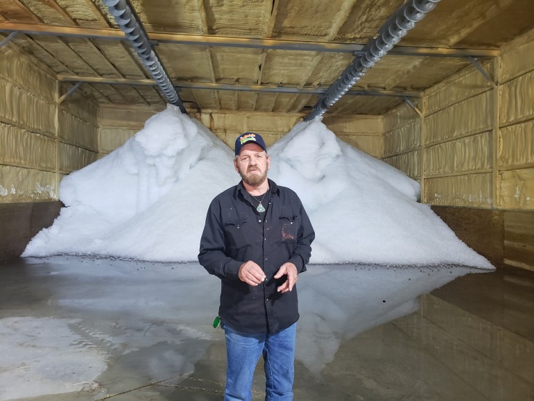 A man in a black ballcap, a black shirt and jeans stands in front of a two giant piles of ice in a warehouse.