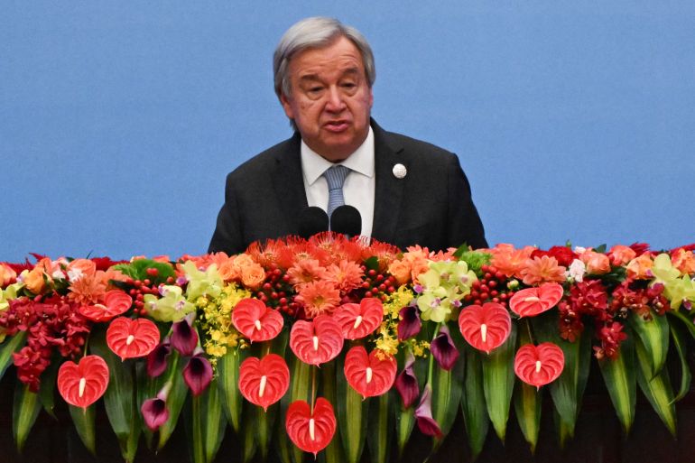 a man in a suit in front of a blue background and a podium of red and yellow flowers