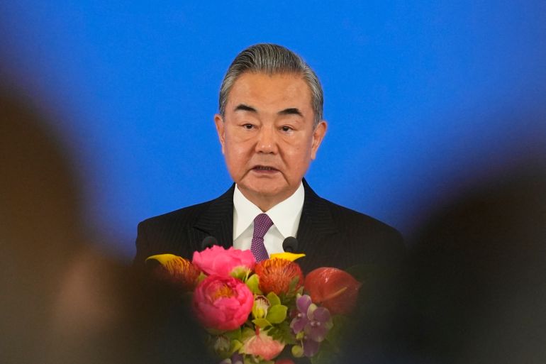 Wang Yi speaking at an event. He has flowers in front of him. The backdrop is blue.