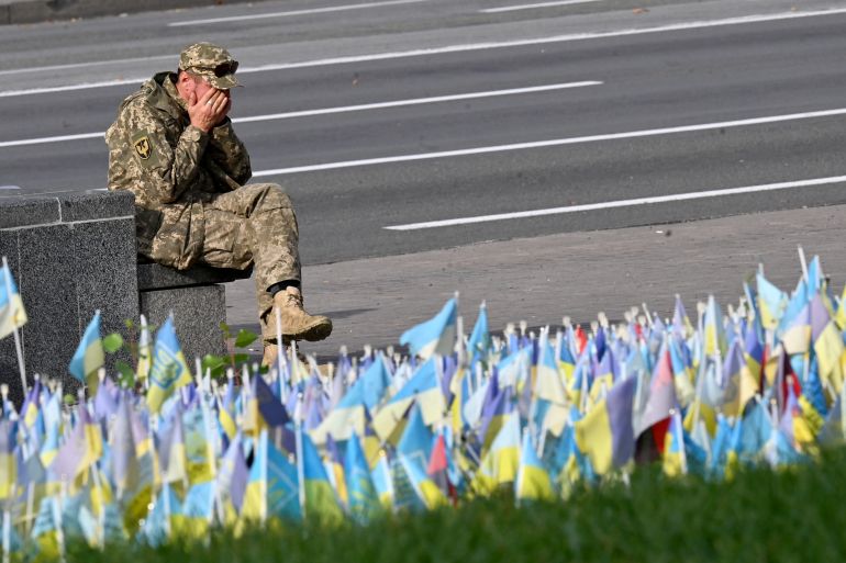 Um militar ucraniano sentado em frente a bandeiras ucranianas que simbolizam soldados ucranianos caídos em um memorial em Kiev.  Ele está com o rosto nas mãos.