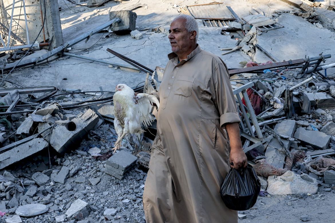A man walks carrying a live chicken past the rubble of a building