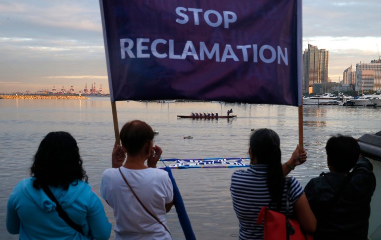 Protesters holding a banner reading 'Stop Reclamation'. They are looking out across the bay. There is a dragon boat crew on the water