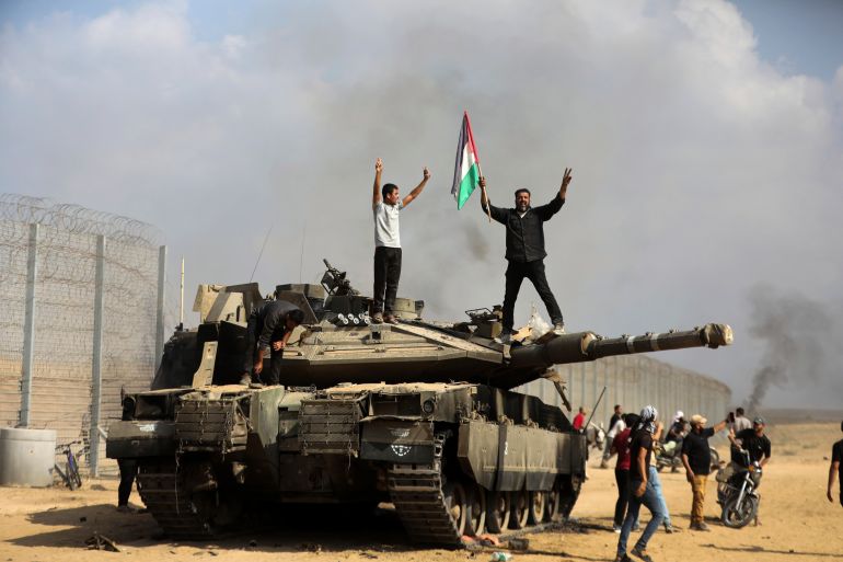 Os palestinos agitam sua bandeira nacional e comemoram perto de um tanque israelense destruído na cerca da Faixa de Gaza, a leste de Khan Younis, no sul, no sábado, 7 de outubro de 2023. Os governantes militantes do Hamas na Faixa de Gaza realizaram um ataque multifrontal sem precedentes contra Israel em ao amanhecer de sábado, disparando milhares de foguetes enquanto dezenas de combatentes do Hamas se infiltravam na fronteira fortemente fortificada em vários locais por via aérea, terrestre e marítima e pegando o país desprevenido em um feriado importante.  (Foto AP/Yousef Masoud)