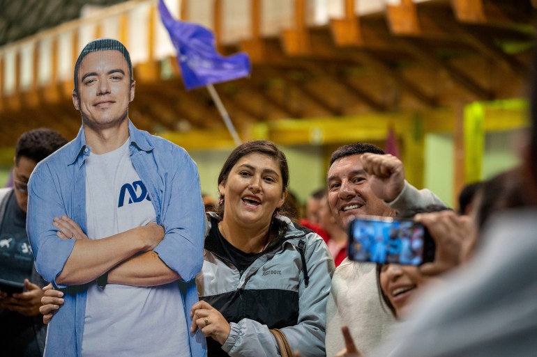 A supporter lifts up a life-sized cardboard picture of Daniel Noboa, while someone else takes a cellphone photo of the scene.