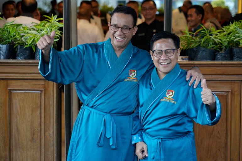 Anies Baswedan, left, and his running mate Muhaimin Iskandar smile at reporters and give a thumbs-up. They are wearing blue robes ahead of a medical check-up required of every candidate