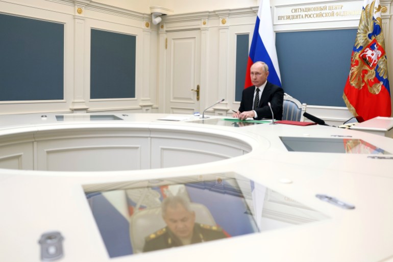 Putin seated at a round table in the Kremlin. The room is white with Russian flags. Defence minister Sergey Shoigu can be seen on a TV screen embedded in the table.