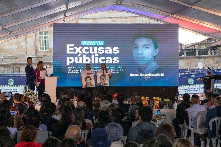 Family members hold up portraits of their loved ones as they remember the 19 victims murdered in extrajudicial killings on October 3 in Colombia. A blue screen behind them reads, "Excusas publicas" and shows the face of one of the victims.