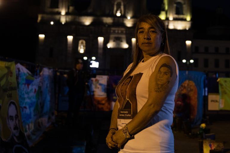 A woman in a white T-shirt stands in a darkened room, where the Colombian government is holding a public event. On her upper arm is a tattoo of a face, representing her 19-year-old son