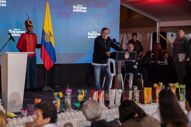 President Gustavo Petro speaks at a podium behind a row of colorful boots, meant to represent 19 people killed by extrajudicial killings. Behind Petro is a blue screen, and a Colombian flag is also visible on stage.