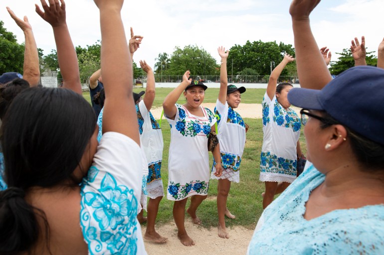 Women dressed in blue-and-white huipil dresses lift their hands as they stand in a circle, doing a team cheer.