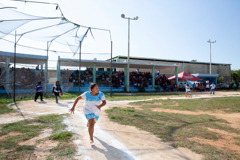 A teenager — barefoot and dressed in a white huipil dress with blue embroidery — runs from home plate to first base at a softball game.