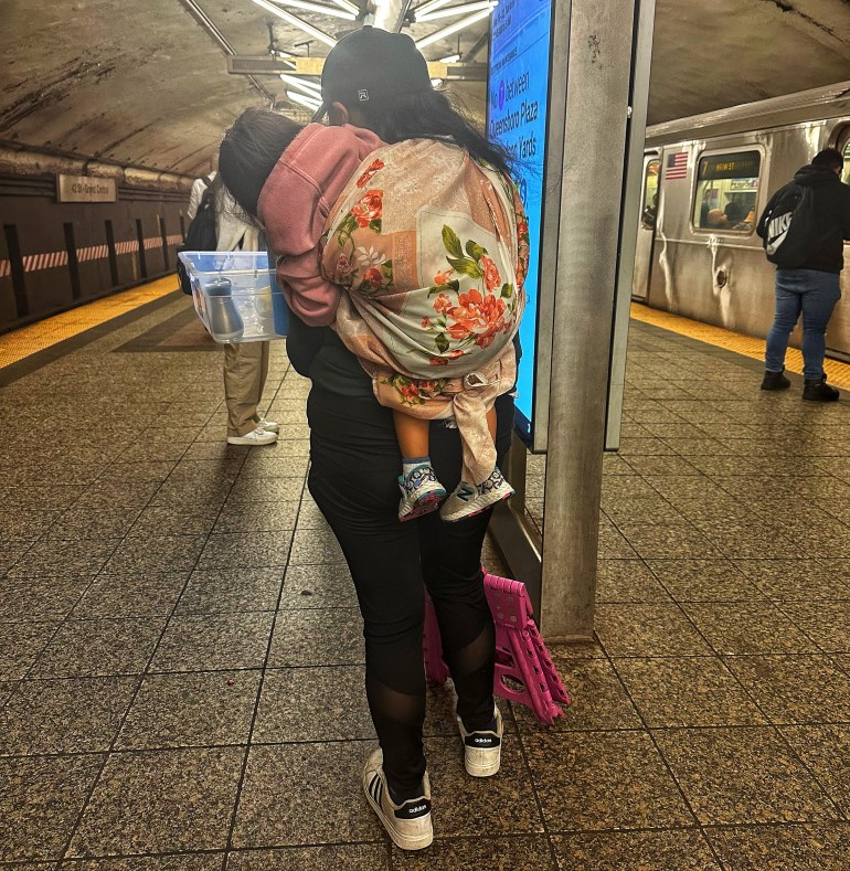 A woman, seen from behind, carries a two-year-old on her back in a quilt through across a subway platform