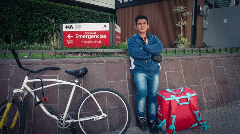 A young man leans against a wall outside a healthcare center, with a sign for "Emergencias." To one side of the man is his bike; to the other is his insulated package for carrying food deliveries.
