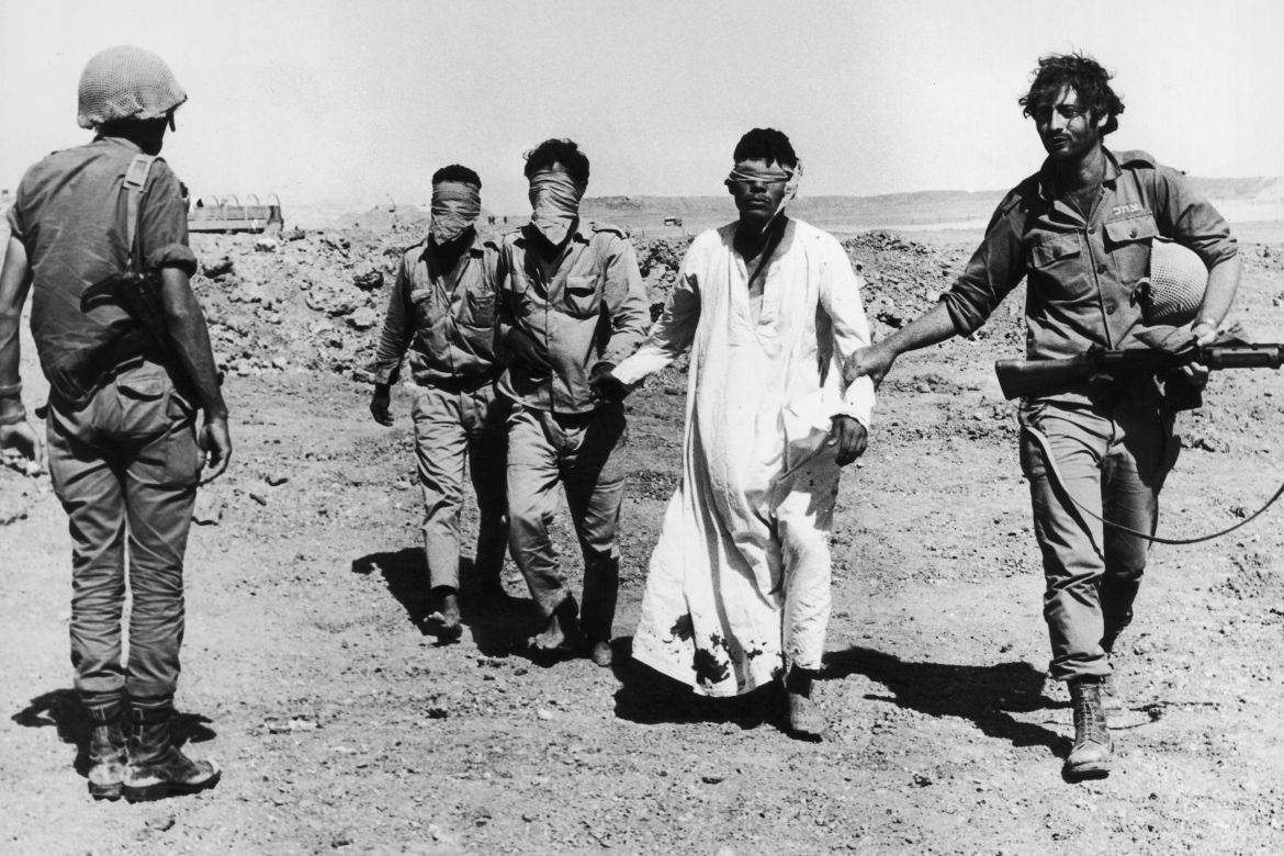 Three barefoot, blindfolded Egyptian POWs are led to the western side of the Suez Canal by an Israeli soldier as another soldier looks on during the Yom Kippur War