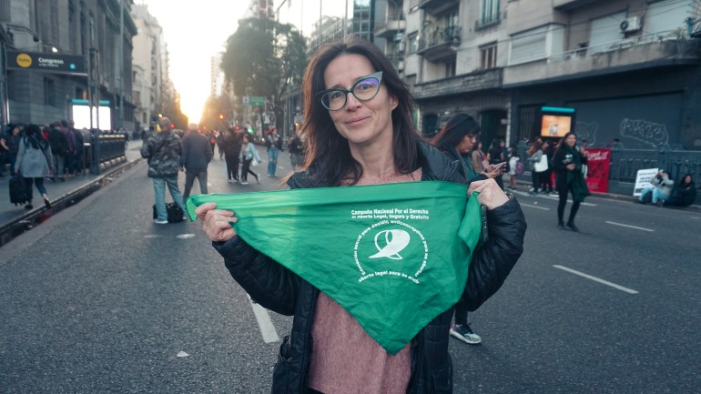 A young woman with glasses standing in a city street holds up a green bandana, symbolising the fight for abortion rights.