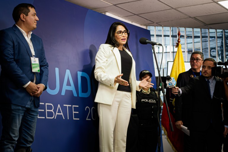 Luisa Gonzalez stands on a debate stage, speaking into a microphone, in front of a banner that reads: Ecuador debate.