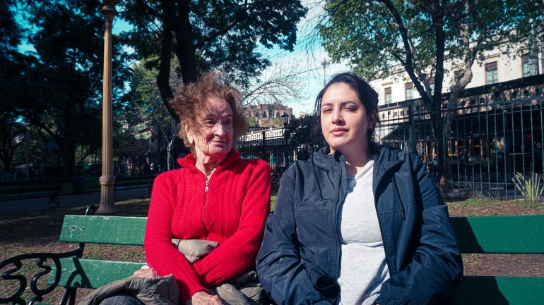 Two women sit side by side on a park bench: one older, one younger.