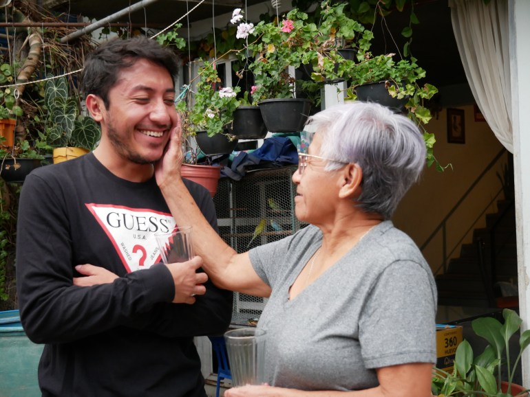 Aunt Paola — a woman with short-cropped grey hair and a grey shirt — lovingly taps the cheek of her grandson Jonathan, who crosses his arms and smiles broadly.
