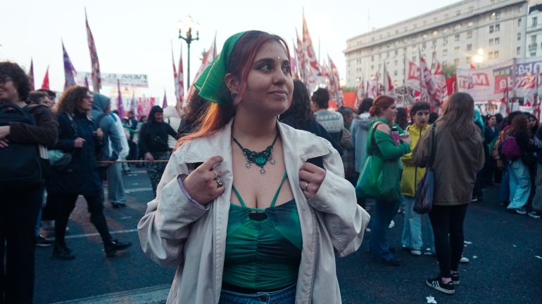 A young woman with a green scarf in her hair — symbolizing the fight for abortion rights — stands in a busy street where a protest is unfolding.