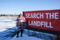 On snow-patched ground outside in Manitoba, a woman — bundled in a white beanie hat and a long plaid coat — stands beside a large red sign that reads, "Search the Landfill."