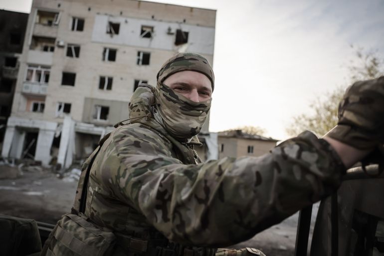 A Ukrainian drone operator on the front line in the Zaporizhia region. He is sitting in an open truck. There are damaged blocks of flats behind him.