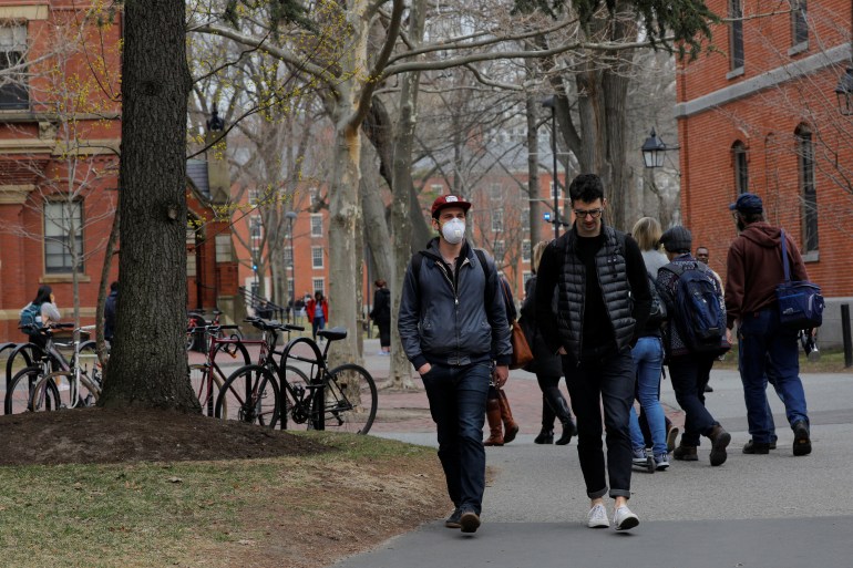 Students in face masks walk across Harvard Yard, brick buildings and a bike rack visible in the background. 