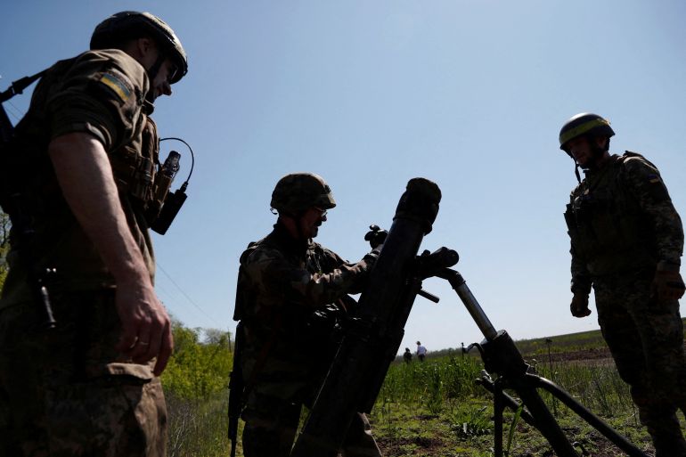 Three members of Ukraine's 128th Mountain Assault Brigade setting up an 120mm mortar during training in May. They are silhouetted against a blue sky. The mortar is in the middle.