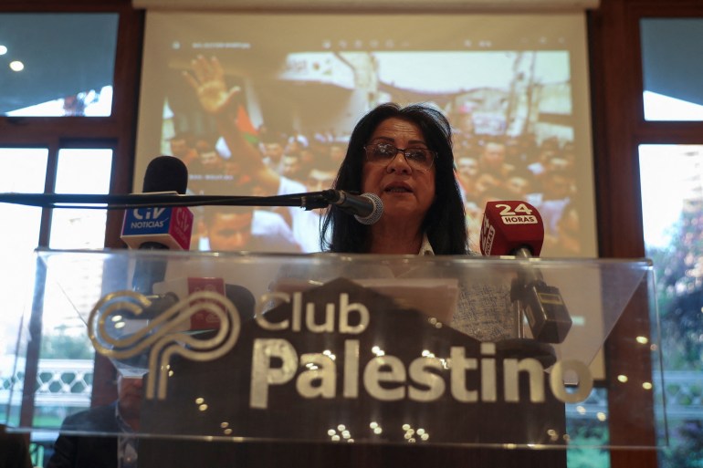 A woman stands in front of a glass podium engraved with the name "Palestinian Club," as she speaks into a microphone.