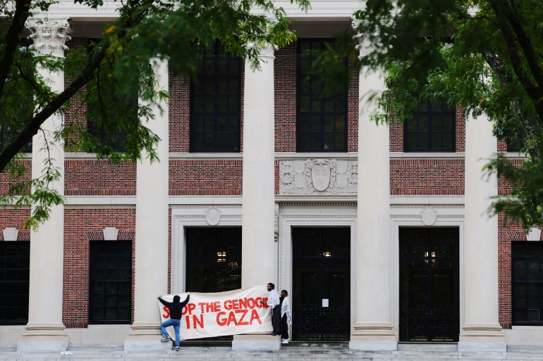 In front of a brick and white-columned building on Harvard's campus, two students raise a banner written in red paint that reads: "Stop the Genocide in Gaza"