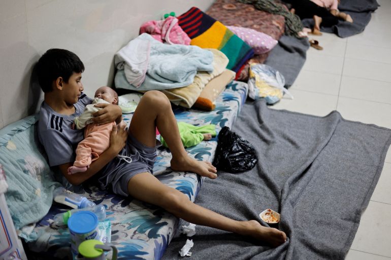 A boy cradles a little baby in his arms as he lies on blankets on the floors of a hospital.