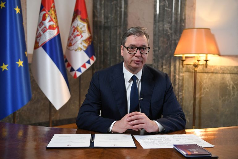 Serbian President Aleksandar Vucic pictired in his office. He is at a desk with papers laid out in front of him. There are flags behind.