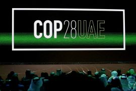 'Cop28 UAE' logo in white is displayed on the screen with a green beam across it, dimly lit audience in the crowd in Abu Dhabi
