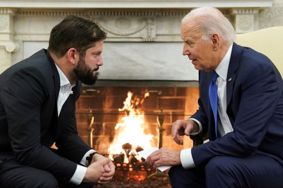 Gabriel Boric and Joe Biden face each other and talk in the Oval Office, a roaring fire in the fireplace behind them.