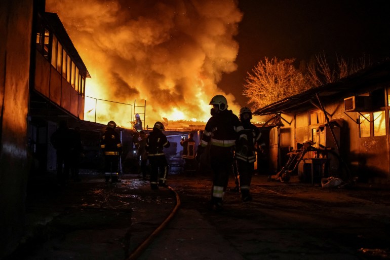 Emergency teams working to put out a fire caused by a Russian drone attack. It's night time. The workers and burning buildings are silhouetted against the orange flames.