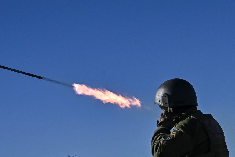 A rocket flies through a blue sky with flames in its wake. A Ukrainian soldier is in the foreground with his hands over his ears.