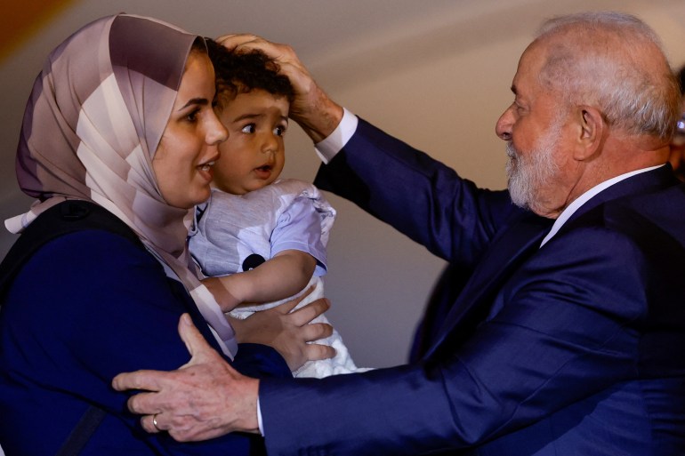 O presidente Luiz Inácio Lula da Silva coloca uma das mãos na cabeça de um menino e outra no braço da mulher que o carrega, como forma de saudação.