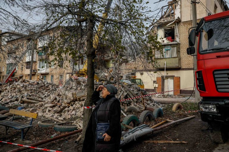 Large piles of rubble from apartment buildings and houses hit by Russian attacks. A woman is in front. A fire engine is also in site.