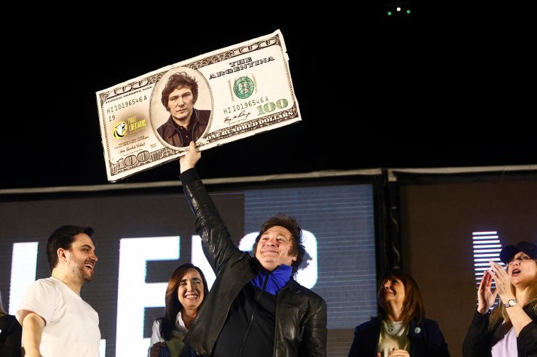 Javier Milei, dressed in a dark suit and dark shirt, holds up an oversized dollar bill with his face in the middle.