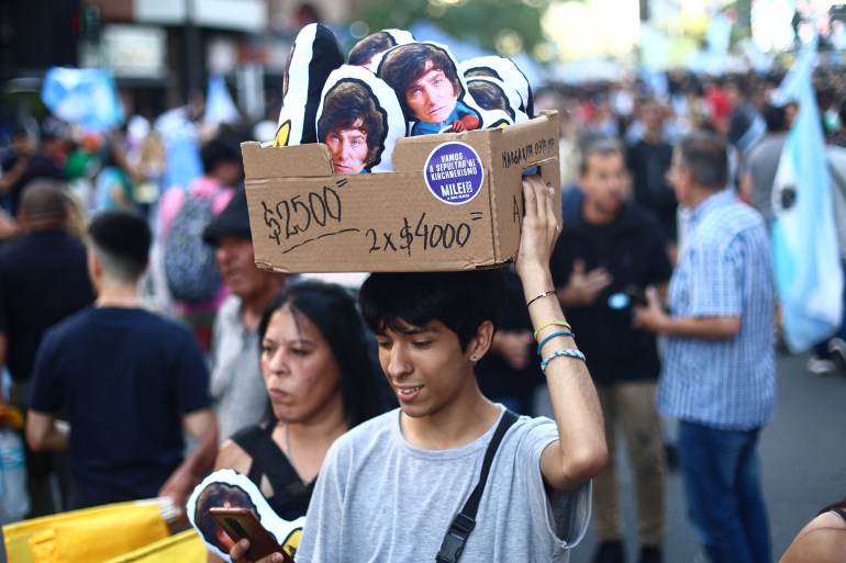 A vendor carries a cardboard box of souvenirs atop his head: They look like stuffed dolls printed with the face of Javier Milei.