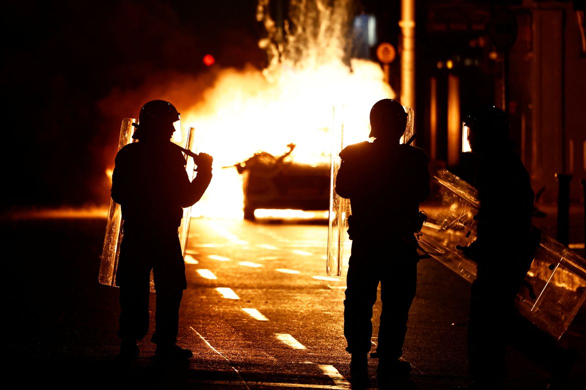 Riot police stands next to a burning police vehicle
