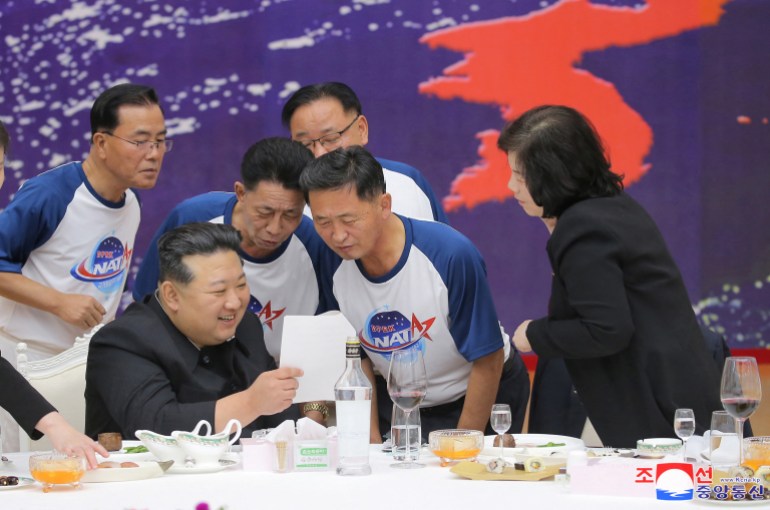 Kim, sentada em uma mesa de banquete, olhando para um pedaço de papel com pessoas vestidas com camisetas NATA.