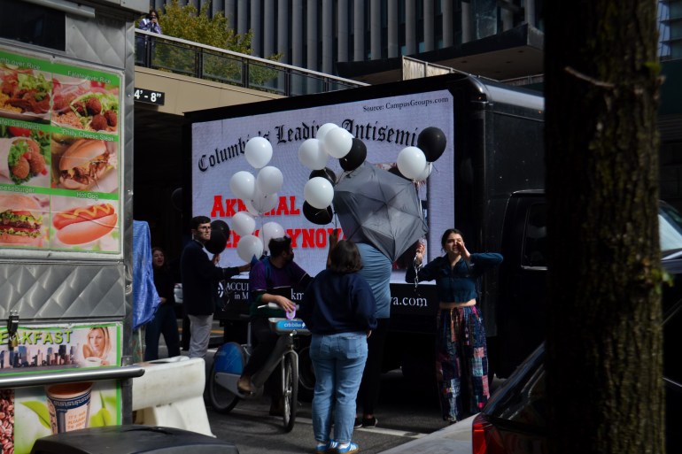 Next to a hotdog stand in New York City, a mobile billboard is parked, advertising students as "Columbia's leading antisemites." But other students stand in front of the mobile billboard, obscuring its message with balloons and umbrellas.