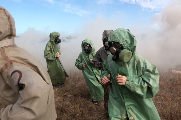 Children in gas masks and green rubber suits. They are running behind a soldier. There is dust and smoke.