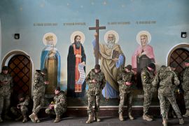Ukrianian soldiers sheltering from the rain in front of a religious fresco. The men are mostly looking at their phones.