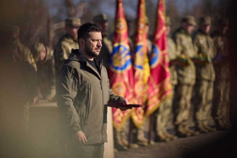 Ukrainian President Volodymyr Zelenskyy. There are soldiers in combat uniform behind him, as well as regimental flags. He is wearing a khaki-coloured jacket.