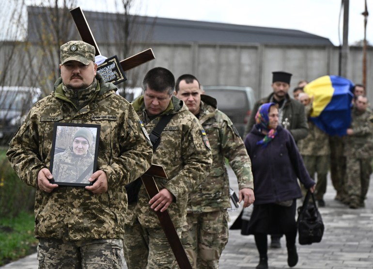 Ukrainian soldiers lead a procession at the funeral of a fellow soldier.  One soldier walks in front with a photo of the dead man, others walk behind with a cross.  A priest walks in front of the coffin, covered with the Ukrainian flag.