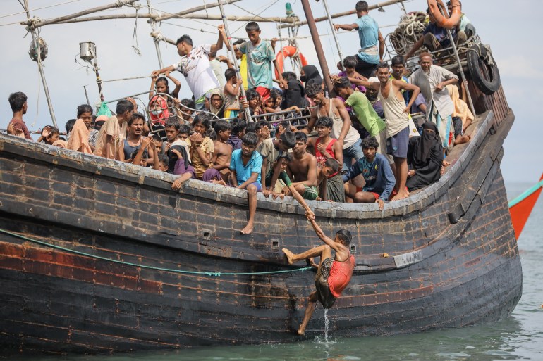 Refugiados Rohingya amontoados no convés de um barco de madeira.  Uma pessoa está subindo pela lateral.