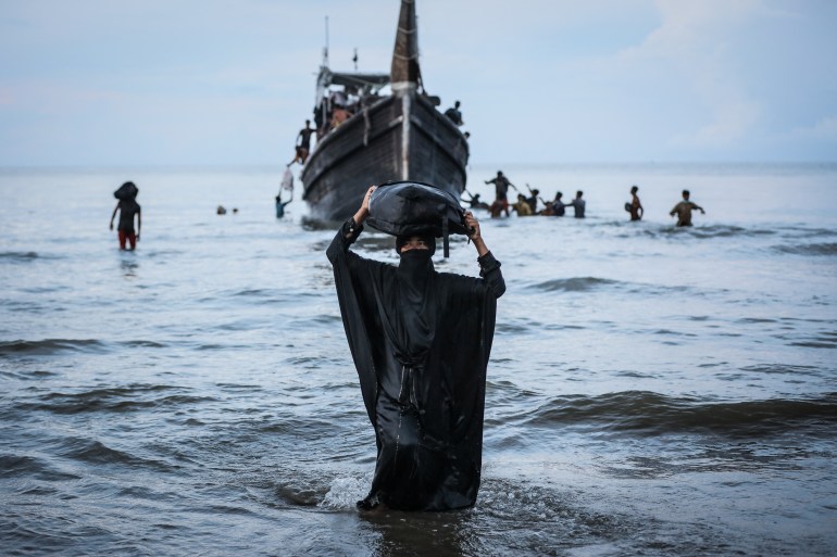 A Rohingya woman in a black robe wading onto the beach. The boat is behind her and she is balancing some belongings on her head